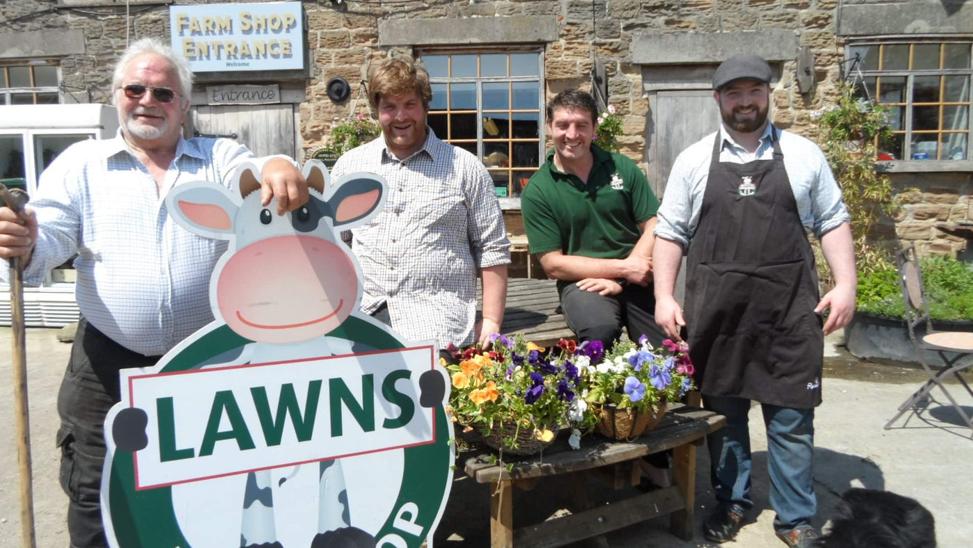 Four men standing in front of a building with a 'Farm Shop Entrance' sign, holding a sign for 'Lawns Farm Shop'.